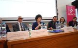 Panelistka przemawia z mikrofonem podczas sesji konferencyjnej przy stole prezydialnym.