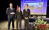 Trzy osoby w eleganckich garniturach pozują na scenie na tle prezentacji „Immatrykulacja studentów”.