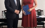 Mężczyzna w garniturze i kobieta w czerwonej sukience pozują z granatową teczką w sali posiedzeń.