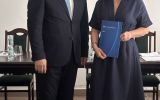 Mężczyzna w garniturze i kobieta w granatowej sukience pozują z granatową teczką w sali posiedzeń.