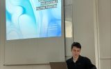Prelegent siedzi przy laptopie i notatkach w sali wykładowej. Na ekranie wyświetlony jest slajd tytułowy prezentacji o podatkach ekologicznych jako narzędziu polityki państwa.