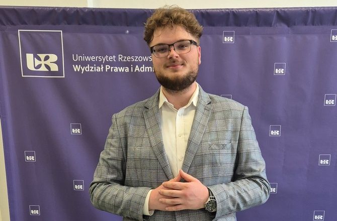 Mężczyzna ubrany w szarą marynarkę w kratę i białą koszulę na tle banera Wydziału Prawa i Administracji Uniwersytetu Rzeszowskiego.
