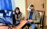 Uczestnik w goglach VR siedzi przy komputerze i trzyma kontrolery. Obok instruktorka prowadzi ćwiczenie z wykorzystaniem symulacji kryminalistycznej.