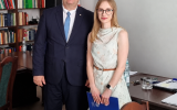 Mężczyzna w garniturze i kobieta w jasnej sukience pozują w sali posiedzeń. Kobieta trzyma granatową teczkę.