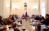 Szerokie ujęcie spotkania w sali konferencyjnej, z uczestnikami siedzącymi po obu stronach długiego stołu.