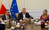 Uczestnicy spotkania siedzą przy stole konferencyjnym obok flag Polski i Unii Europejskiej, rozmawiając w formalnej sali.