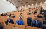 Uczestnicy konferencji siedzą w dużej sali audytoryjnej i słuchają wystąpienia. Widownia zajmuje kilka rzędów miejsc.
