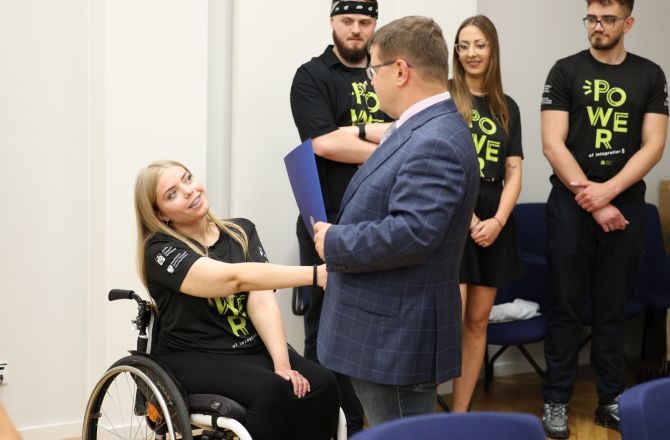 Zawodniczka na wózku inwalidzkim otrzymuje list gratulacyjny od rektora UR. Z tyłu trenerzy oraz inni sportowcy ubrani w klubowe, czarne koszulki z żółtymi napisami "POWER"