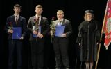 Trzech studentów trzymających niebieskie teczki i czerwone róże pozuje do zdjęcia z przedstawicielką wydziału w stroju akademickim.