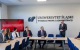 Uczestnicy spotkania siedzą przy stole konferencyjnym na Wydziale Prawa i Administracji Uniwersytetu Śląskiego. Jeden z mężczyzn stoi i przemawia, a w tle widoczny jest ekran z napisem „Konferencja Dziekanów Publicznych Uniwersyteckich Wydziałów Prawa”.