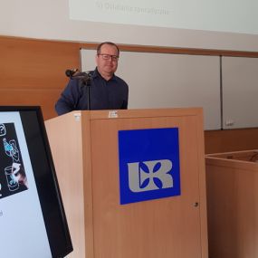 Za mównicą dr Sławomir Dybka. Z przodu logo Uniwersytetu Rzeszowskiego. W tle widok slajdu dotyczącego prowadzonego warsztatu.