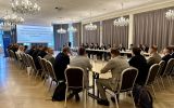 Uczestnicy konferencji siedzą przy stołach ustawionych w podkowę i słuchają wystąpienia.
