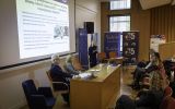 Prelegentka prowadzi wystąpienie przy ekranie prezentacyjnym; uczestnicy siedzą w auli.