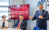 Mężczyzna w garniturze wypowiada się podczas spotkania konferencyjnego. Obok niego przy stole siedzą uczestnicy, a w tle znajduje się ekran z napisem „Konferencja Dziekanów Publicznych Uniwersyteckich Wydziałów Prawa”.