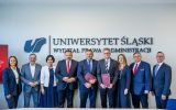Grupa uczestników spotkania pozuje do wspólnego zdjęcia na Wydziale Prawa i Administracji Uniwersytetu Śląskiego. W tle widoczne jest logo i nazwa uczelni, a dwie osoby trzymają bordowe teczki.