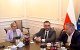 Troje uczestników spotkania siedzi przy stole konferencyjnym podczas rozmowy w sali z flagami Polski i Unii Europejskiej.