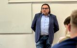 Prelegent stoi w sali dydaktycznej przed tablicą i prowadzi spotkanie ze studentami.