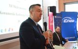Prelegent w garniturze przemawia z mikrofonem na tle ekranu konferencyjnego i banerów partnerów.