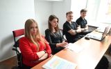 Grupa studentów siedzących przy stołach podczas seminarium. Na pierwszym planie uczestniczka w czerwonej bluzce, obok niej osoby z notatkami i laptopami.
