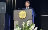 Młody mężczyzna w garniturze przemawia przy mównicy podczas uroczystości akademickiej.