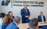 Mężczyzna w garniturze przemawia podczas spotkania na Wydziale Prawa i Administracji Uniwersytetu Śląskiego. Uczestnicy siedzą przy stole konferencyjnym, a w tle widoczne jest logo uczelni.