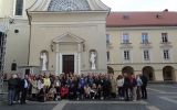 Zdjęcie grupowe uczestników konferencji na Wielkim Dziedzińcu Uniwersytetu Wileńskiego. Kilkadziesiąt osób stoi przed klasycystycznym, jasnym budynkiem uczelni z charakterystycznym frontonem, na którym widnieje łaciński napis „ALMA MATER VILNENSIS”
