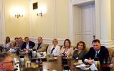 Uczestnicy spotkania siedzą przy stole konferencyjnym i słuchają rozmowy prowadzonej po przeciwnej stronie sali.