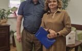 Mężczyzna w ciemnej koszuli i brązowych spodniach oraz kobieta w brązowej sukience koszulowej trzymającą niebieską teczkę stojący w jasnym biurze