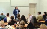 Prelegent przemawia do studentów siedzących w sali dydaktycznej. W tle widoczna jest tablica i ekran projekcyjny.