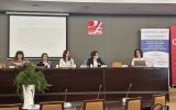 Uczestniczki panelu siedzą przy stole prezydialnym podczas obrad konferencji. W tle widoczny jest ekran projekcyjny oraz roll-up z nazwą konferencji.