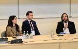 Trzy osoby siedzące przy stole prezydialnym. Mężczyzna z mikrofonem wypowiada się,