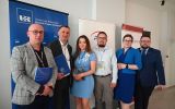 Grupa uczestników konferencji pozuje do zdjęcia na tle banerów organizatorów i partnerów wydarzenia.