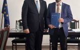 Dwóch mężczyzn pozuje w sali posiedzeń; jeden trzyma granatową teczkę, a obok widoczna jest flaga Unii Europejskiej.