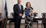 Mężczyzna w garniturze i kobieta w czarno-białej sukience stoją w centrum sali posiedzeń; kobieta trzyma granatową teczkę.