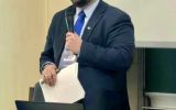 Prelegent w garniturze przemawia z mikrofonem podczas konferencji. Na stole przed nim stoi laptop.