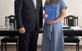 Mężczyzna w garniturze i kobieta w niebieskiej sukience pozują z granatową teczką w sali posiedzeń.