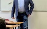 Prelegent stoi w sali dydaktycznej przed tablicą i prowadzi spotkanie ze studentami.