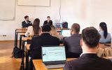 Uczestnicy spotkania siedzą w sali dydaktycznej, obserwując osoby prowadzące prezentację przy stole prezydialnym.