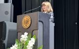 Kobieta w okularach i ciemnej marynarce wygłasza przemówienie przy mównicy podczas uroczystości akademickiej.