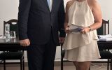 Mężczyzna w garniturze i kobieta w jasnej sukience pozują w sali posiedzeń podczas wręczenia dokumentu.