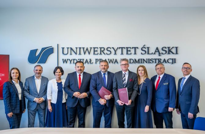 Grupa uczestników spotkania pozuje do wspólnego zdjęcia na Wydziale Prawa i Administracji Uniwersytetu Śląskiego. W tle widoczne jest logo i nazwa uczelni, a dwie osoby trzymają bordowe teczki.