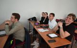 Studenci siedzący w sali wykładowej przy ławkach. Na stolikach leżą notatki, tablety i laptopy.