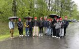 Grupa studentów stojących na zewnątrz, pada deszcz,  w rękach trzymają parasole
