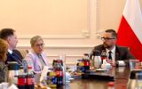 Uczestnicy spotkania rozmawiają przy stole konferencyjnym, w tle widoczna jest flaga Polski.
