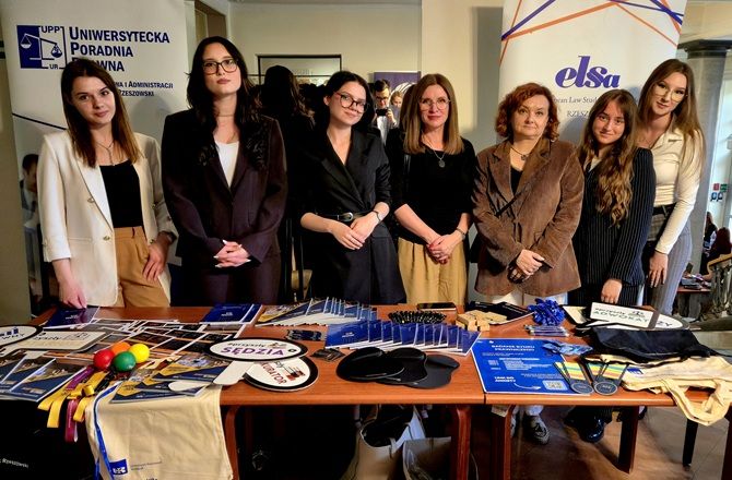 Grupa osób stoi przy stoisku Uniwersyteckiej Poradni Prawnej i ELSA Rzeszów. Na stole znajdują się materiały promocyjne i gadżety edukacyjne.