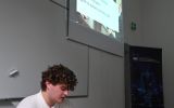 Student prowadzi prezentację przy laptopie w sali dydaktycznej. Na ekranie wyświetlony jest slajd dotyczący kompetencji rady nadzorczej.