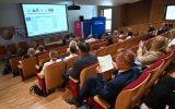 Widok z góry na salę konferencyjną z uczestnikami, ekranem prezentacyjnym i banerami partnerów.