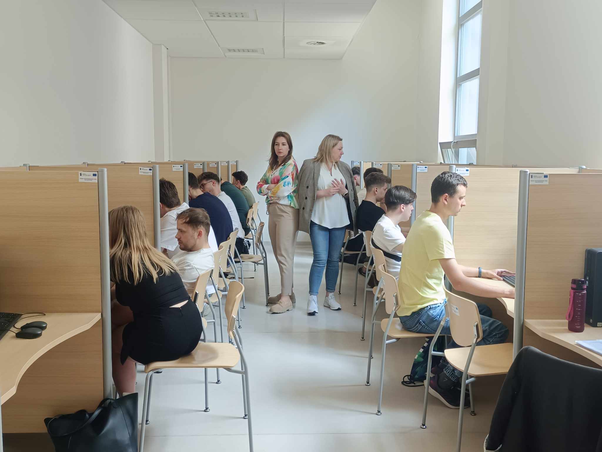 Studenci siedzą przy komputerach, dwoje prowadzących stoi