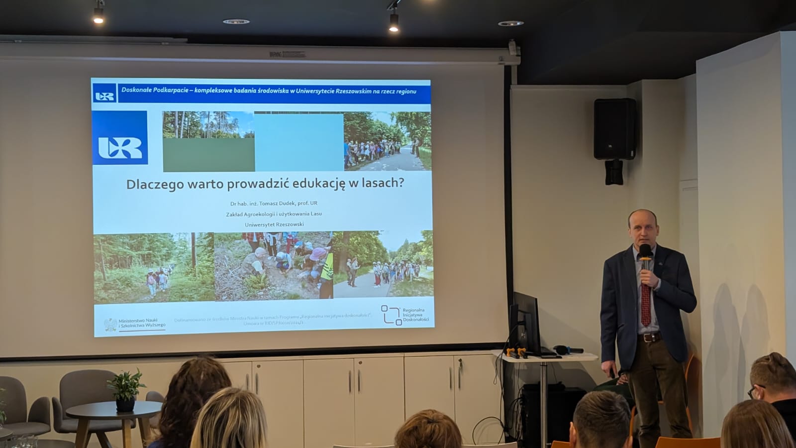 Prof dudek stoi, trzyma w ręku mikrofon i omawia treść slajdu pt "dlaczego warto prowadzić edukację w lasach ?"