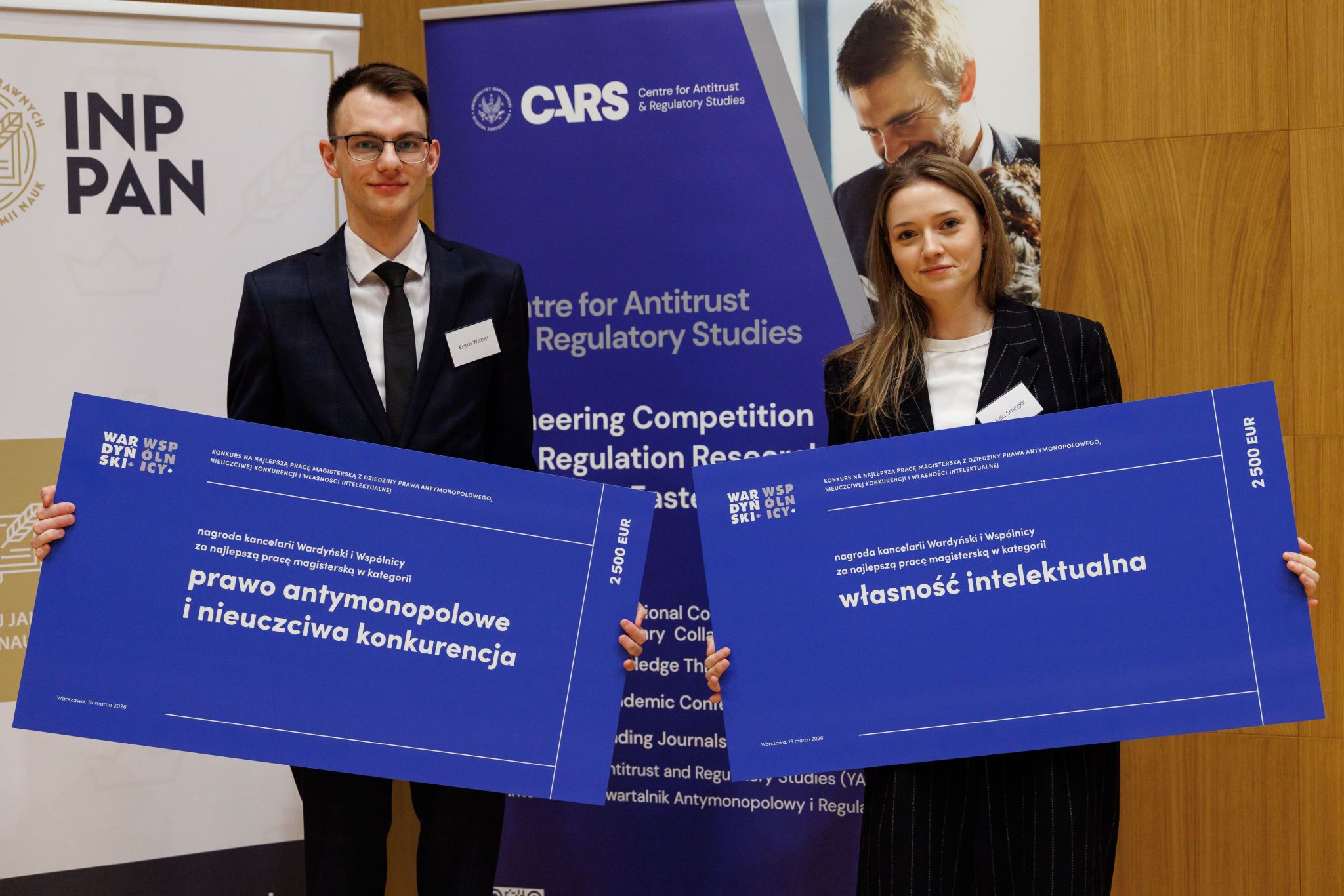Laureaci konkursu na najlepszą pracę magisterską organizowanego przez kancelarię Wardyński i Wsp&oacute;lnicy. Dwie osoby trzymające banery z napisami "prawo antymonopolowe i nieuczciwa konkurencja" oraz "własność intelektualna"